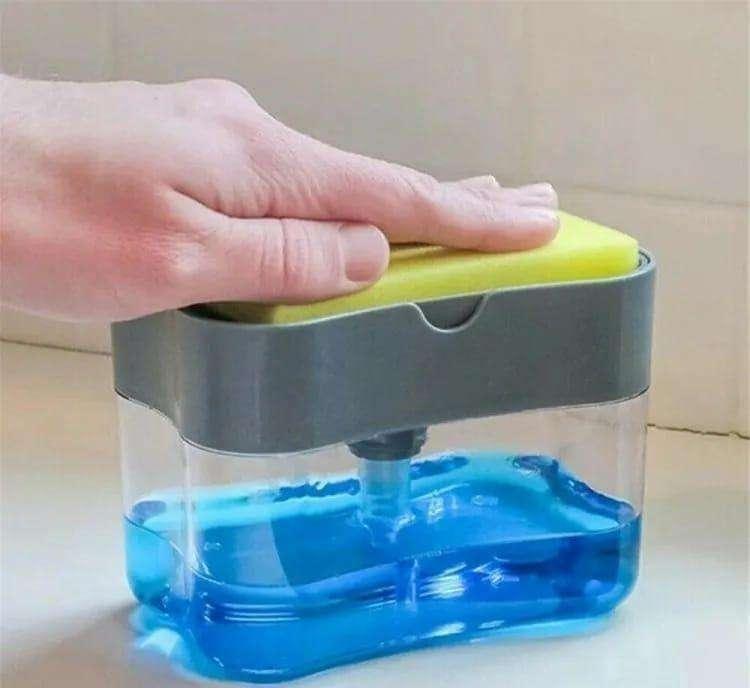 Plastic soap dispenser with gray top and clear body filled with blue soap, hand pressing yellow sponge on top for dispensing liquid soap